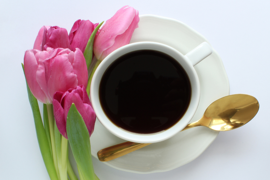 Stock image of a cup of coffee, with a gold spoon and pink tulips resting on saucer
